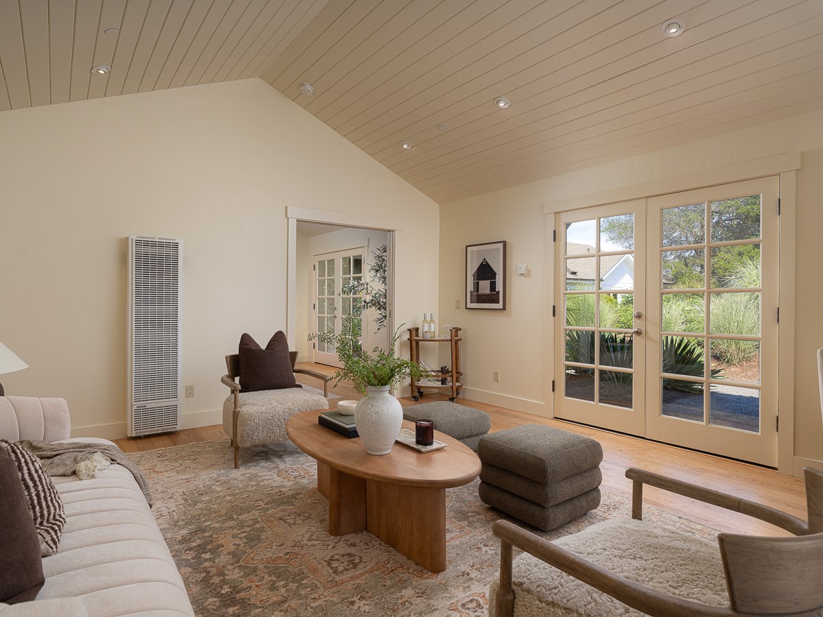 Living room with beige sofa, armchair, wooden coffee table with a vase of green plants, ottomans, and glass double doors leading to outdoor garden.