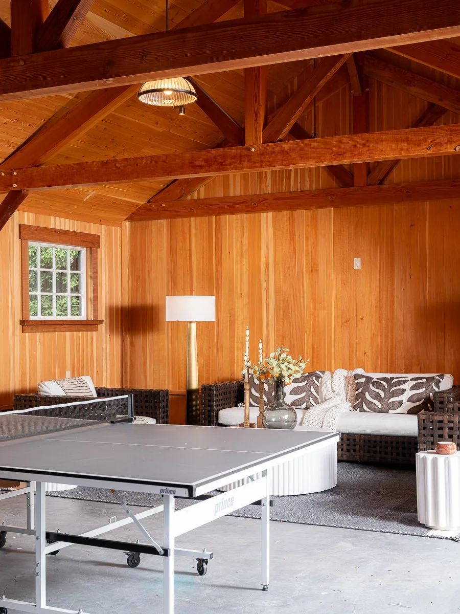 A room with wooden walls and ceiling, featuring a ping pong table and a sitting area with a wicker sofa and a lamp.