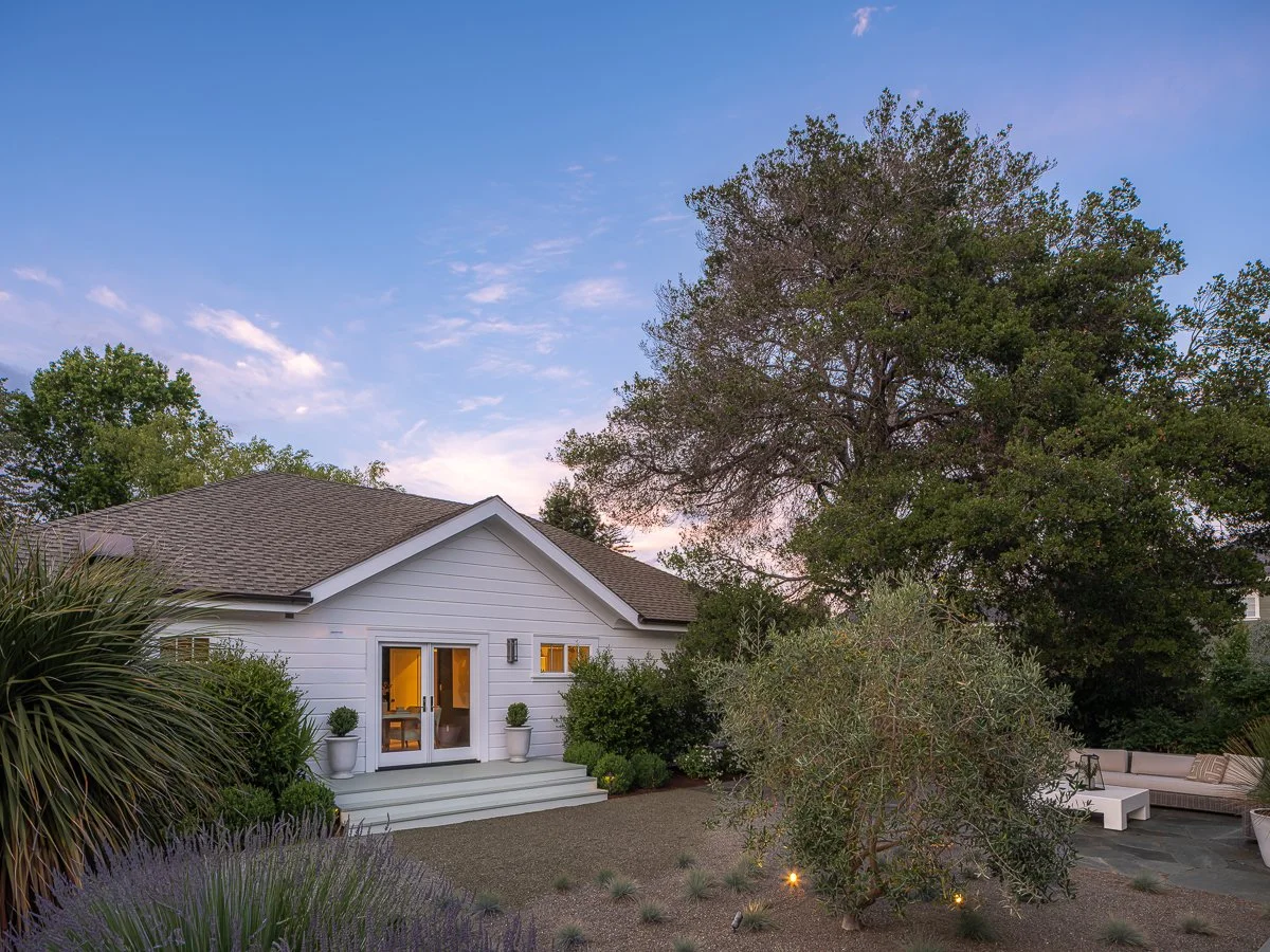 A house with white siding and a grey roof, surrounded by trees and plants, with outdoor seating in a backyard during twilight.
