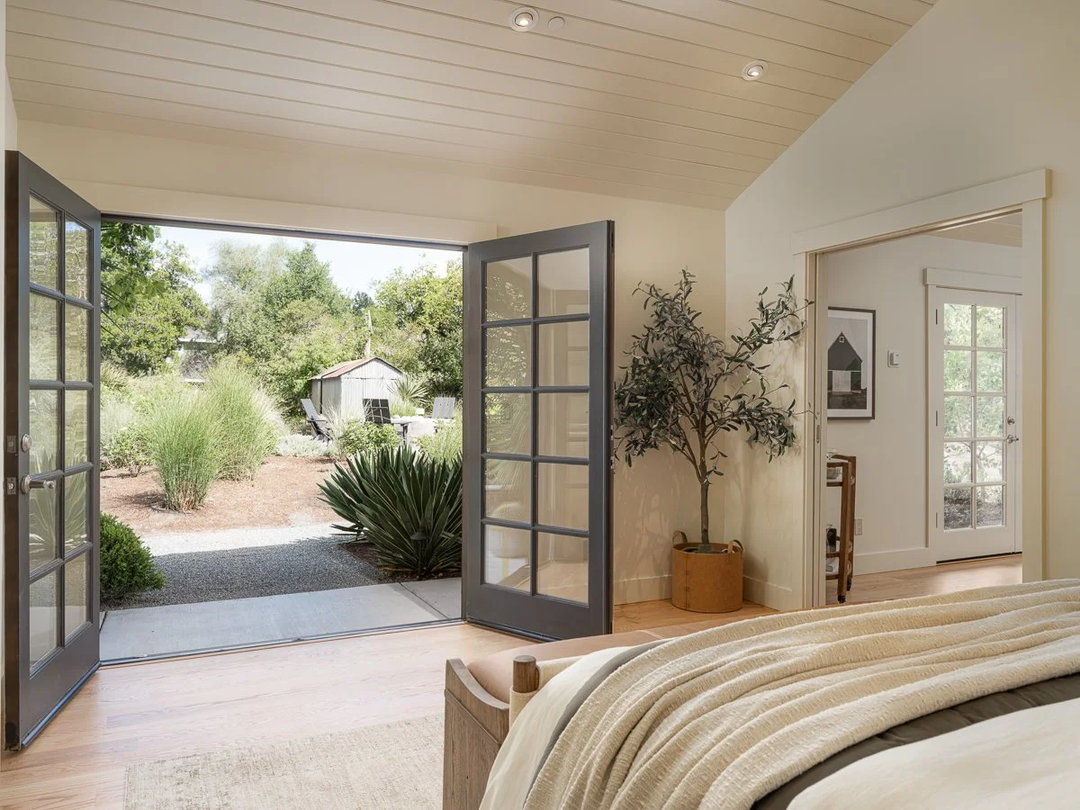 View from a bedroom looking outside through open French doors into a garden with trees, bushes, and outdoor furniture.