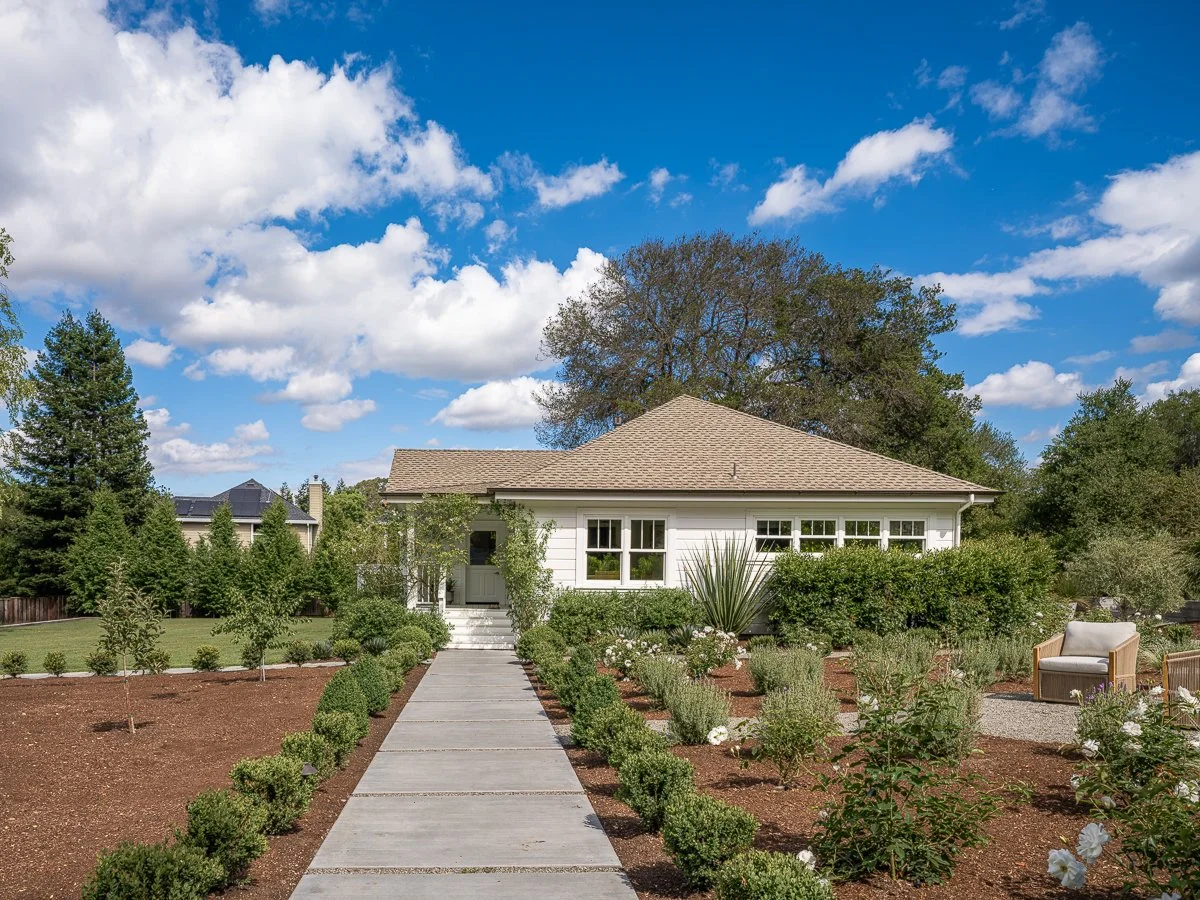 A white house with a gabled roof surrounded by a garden with bushes and trees, and a courtyard with a concrete pathway and outdoor seating, under a blue sky with scattered clouds.