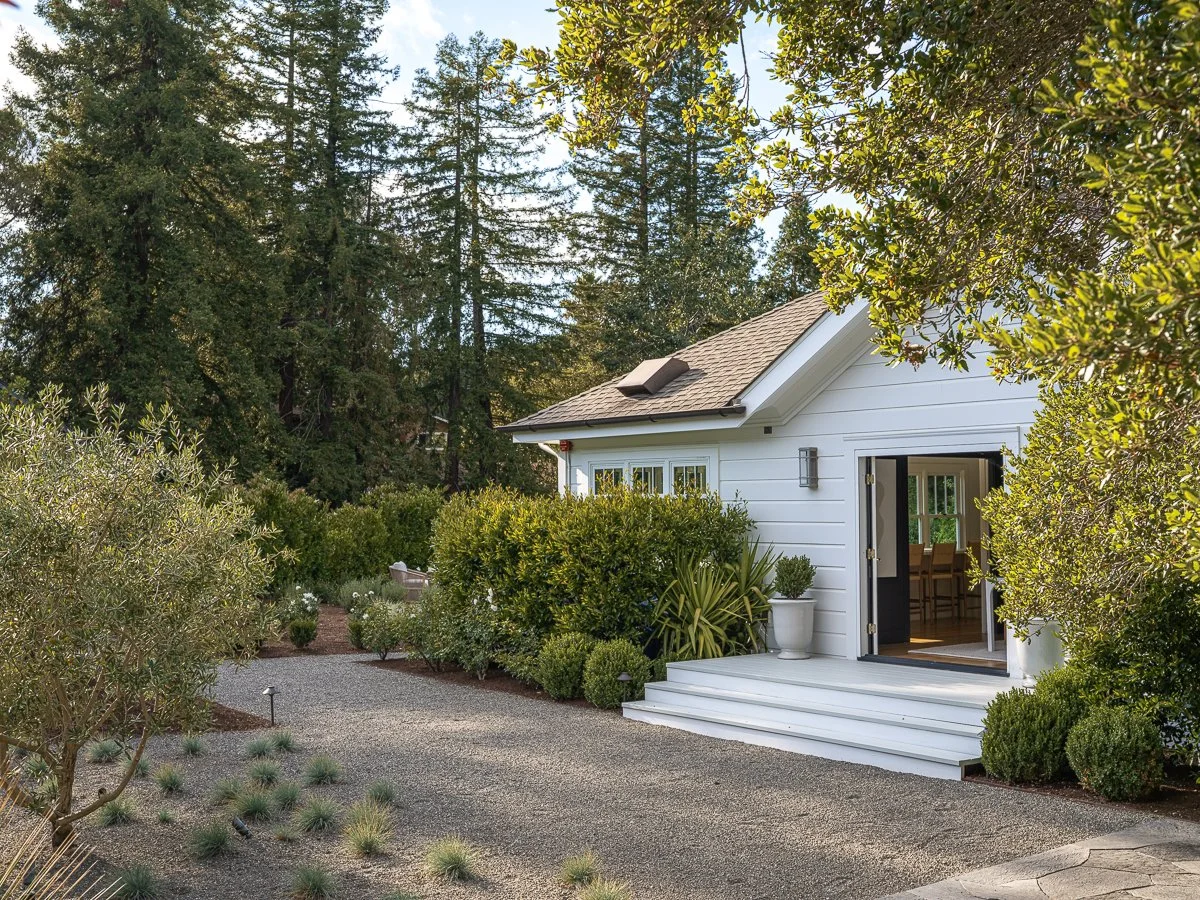 A white house with an open door, surrounded by greenery and trees, with a gravel pathway leading to it.