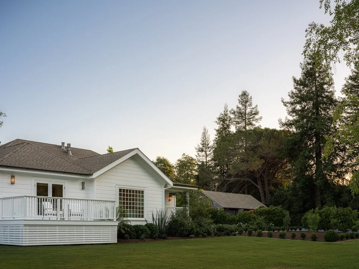 A white house with a wooden deck and attached stairs, surrounded by well-maintained garden and trees, under a clear sky.