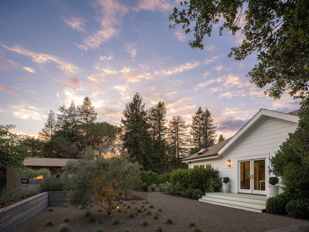 A backyard garden with a white house, potted plants, trees, and a sunset sky with clouds.