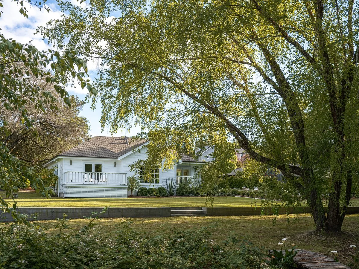 A white house with a deck is seen through large trees in a backyard with a grassy lawn and small plants in the foreground.