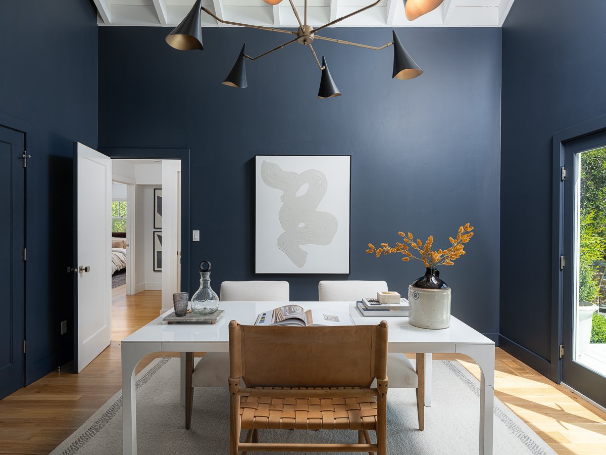 A modern dining room with dark blue walls, a white table, and chairs. There is a piece of abstract art on the wall, a vase with yellow-orange leaves, and a dropper on the table. Sunlight is coming through a glass door.
