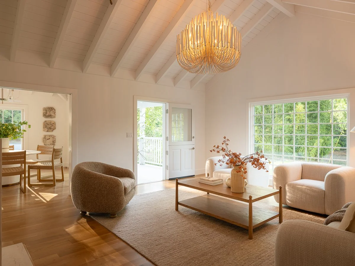 Bright living room with white walls, high vaulted ceiling, a large chandelier, large window, beige sofas, wooden coffee table with vases and books, and a neutral area rug. Adjacent dining area with wooden table and chairs is visible.