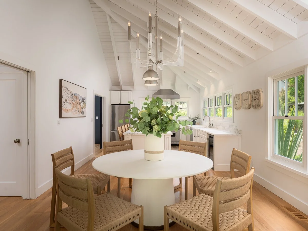 Bright dining area in a modern kitchen with a white round table, six beige woven chairs, a large green plant centerpiece, and a white vaulted ceiling with exposed beams. Large windows provide natural light and greenery views.