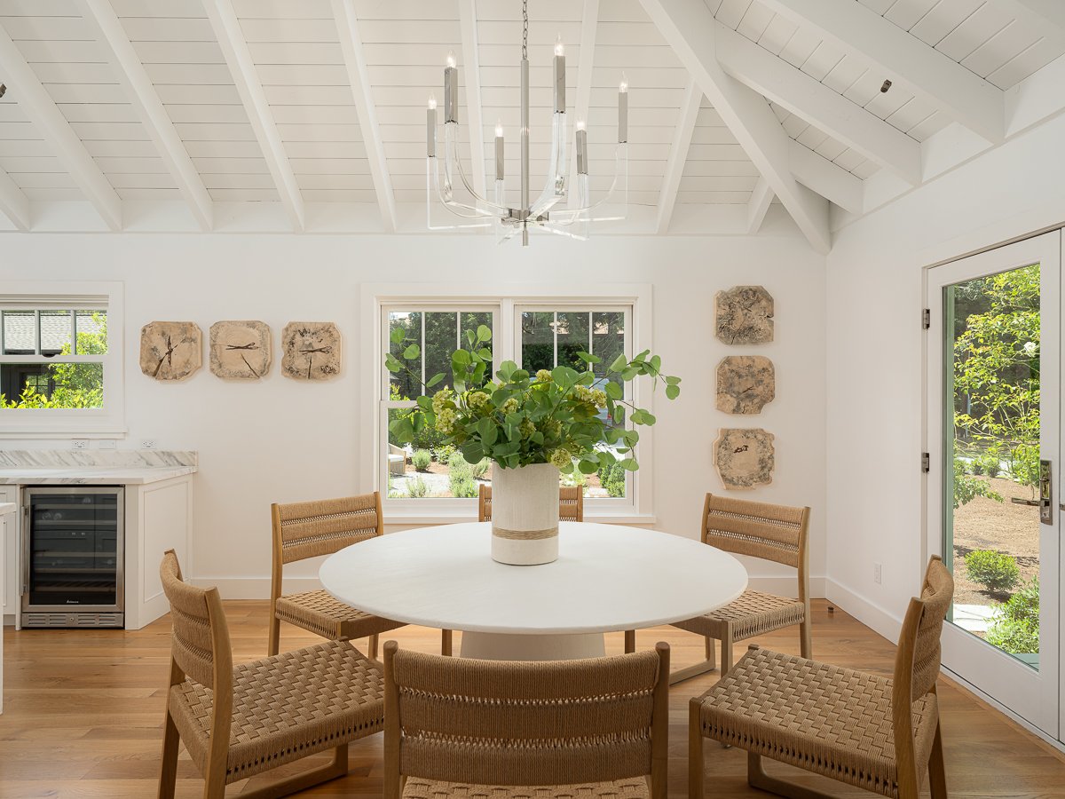A dining room with a white round table, six woven chairs, a floral centerpiece, and white walls with decorative stone art pieces. There are windows, a glass door, and a chandelier hanging from the vaulted ceiling.