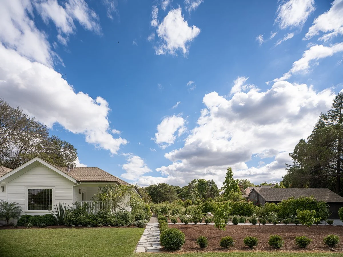 A suburban yard with a white house to the left, a stone pathway, well-maintained green grass, neatly trimmed bushes, and trees under a partly cloudy blue sky.