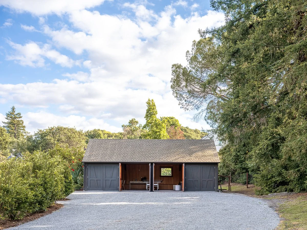 A black garage with open doors showing interior work space, situated on a gravel driveway with trees and bushes surrounding it under a partly cloudy sky.