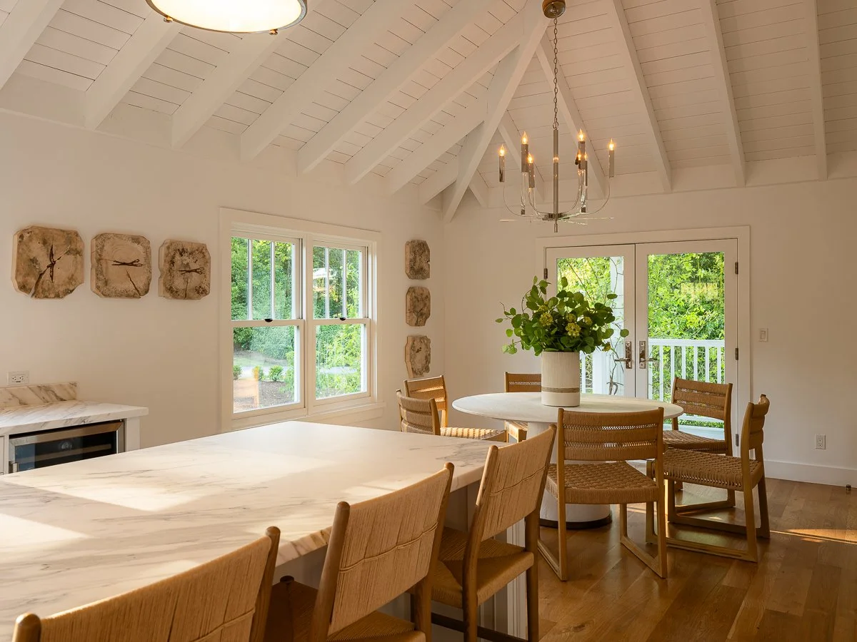Bright dining room with white vaulted ceiling, chandelier, large window, glass doors with greenery outside, round table with a plant centerpiece, wooden chairs, and a partial view of a marble-topped counter.