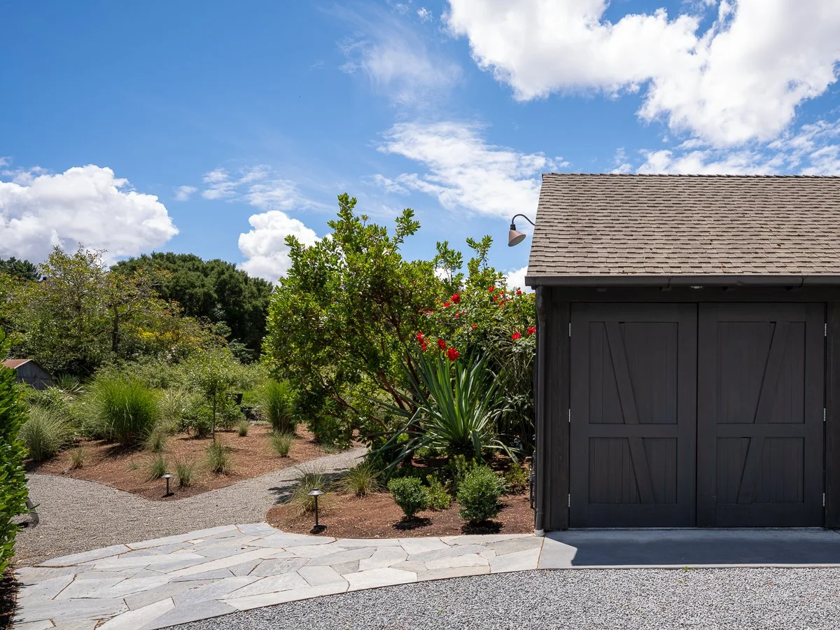 A garden with various plants, a small path, and a dark gray shed with closed double doors under a partly cloudy sky.