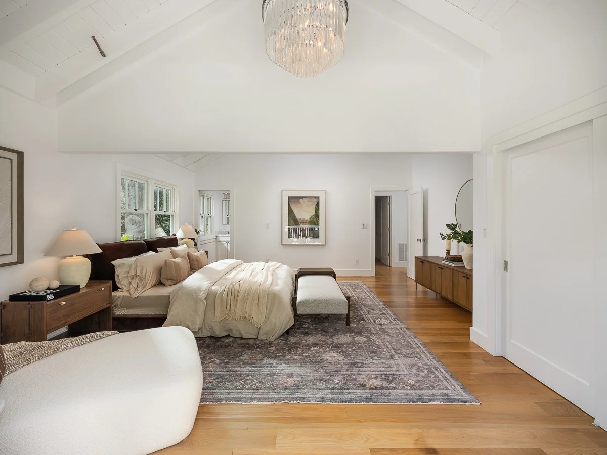 A spacious bedroom with a large bed, wooden furniture, and hardwood floors. Light-colored walls, a crystal chandelier, and windows letting in natural light.
