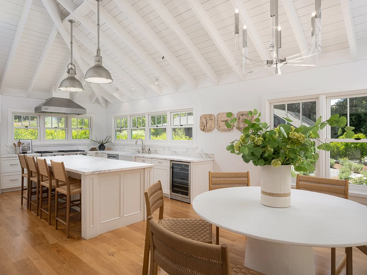 Bright white kitchen with a vaulted ceiling, large windows, wooden floors, a kitchen island with bar stools, and a round dining table with a flower vase.