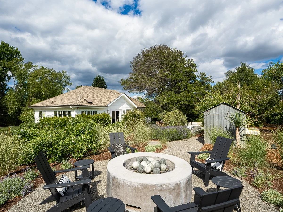 An outdoor patio area with five black Adirondack chairs arranged around a large concrete fire pit filled with decorative stones. The area is landscaped with various plants and shrubs, with a white house and a small metal shed in the background, under