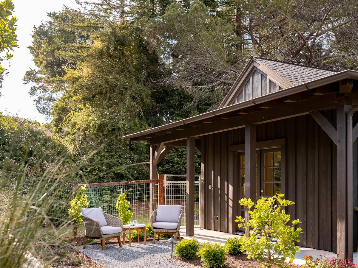 A cozy outdoor seating area with two cushioned chairs and a small table on a gravel surface, in front of a dark wooden house with a sloped roof, surrounded by greenery and trees.