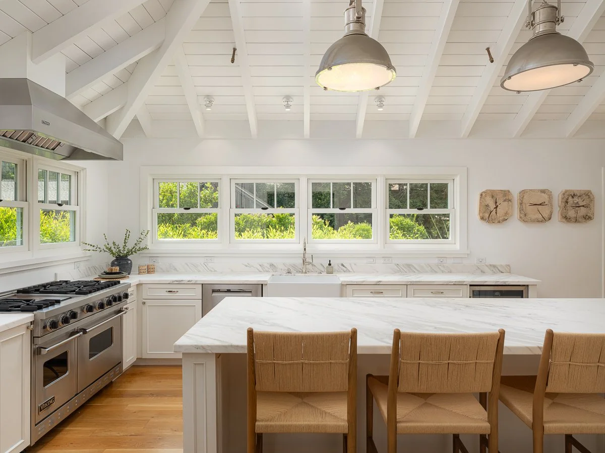 Modern kitchen with white cabinets, marble countertops, and a large island with seating. Stainless steel appliances, multiple windows letting in natural light, and decorative wall art.