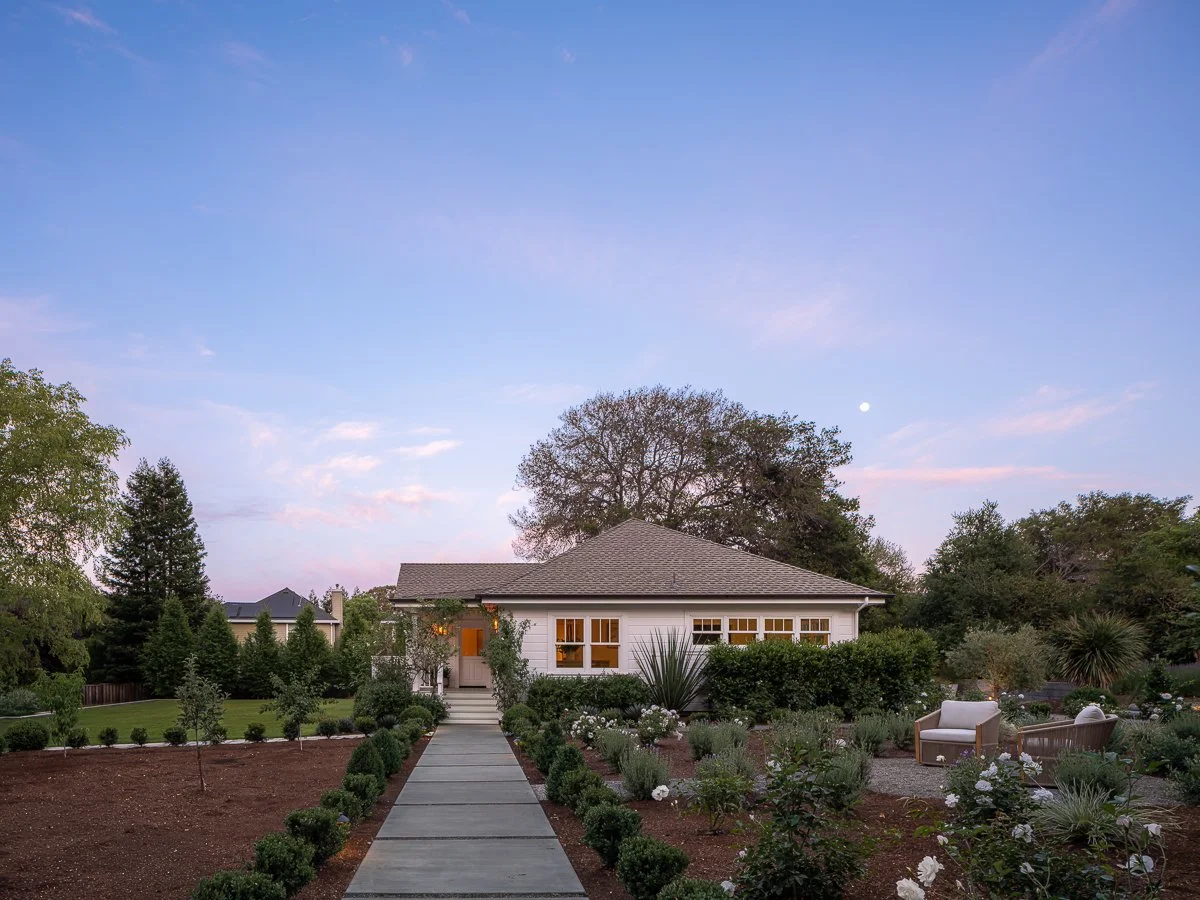 A house with a garden path leading to the front door, surrounded by lush greenery and outdoor furniture, under a sky with a visible moon.