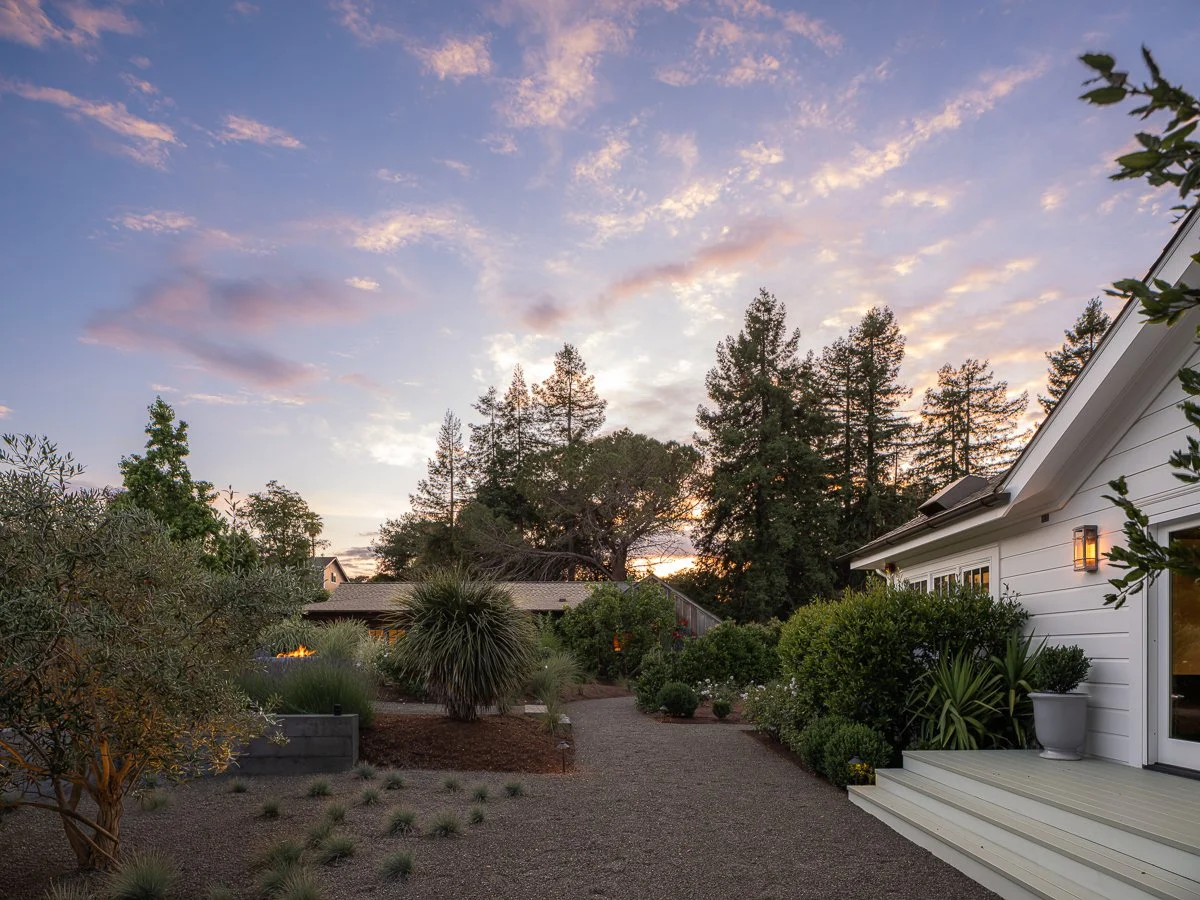 A backyard garden during sunset with a gravel pathway, a white house on the right with a small porch, and trees and shrubs surrounding the area