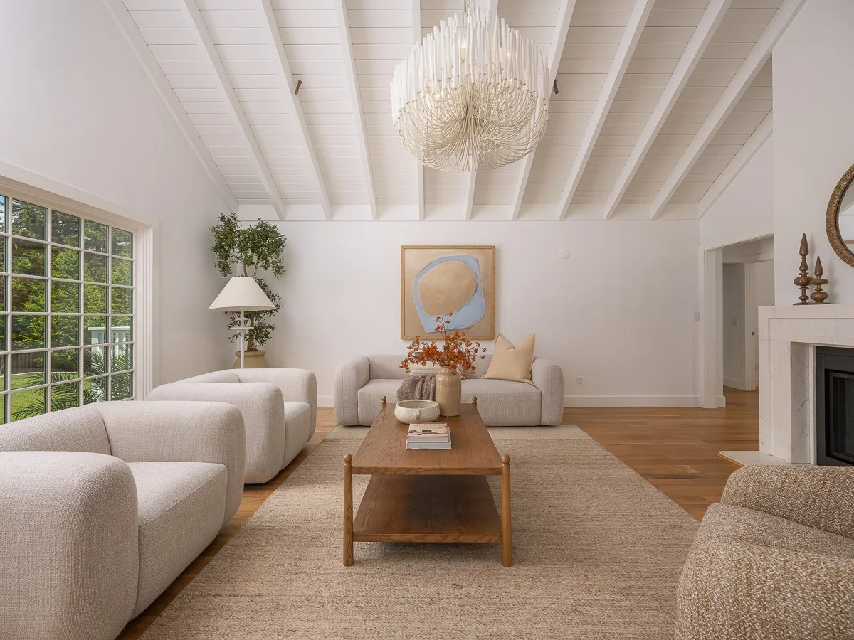 Living room with white walls and vaulted ceiling with wooden beams, large window showing greenery outside, white sofas, a wooden coffee table with decor, a modern chandelier, and a fireplace with decorative items.