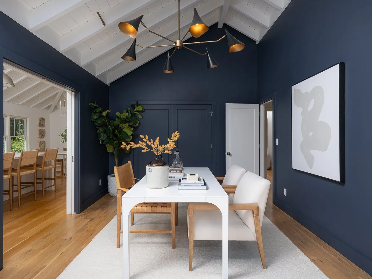 Modern dining room with white table, white and wooden chairs, navy blue walls, large black and gold chandelier, indoor plant, abstract wall art, and access to a light-filled adjacent room.