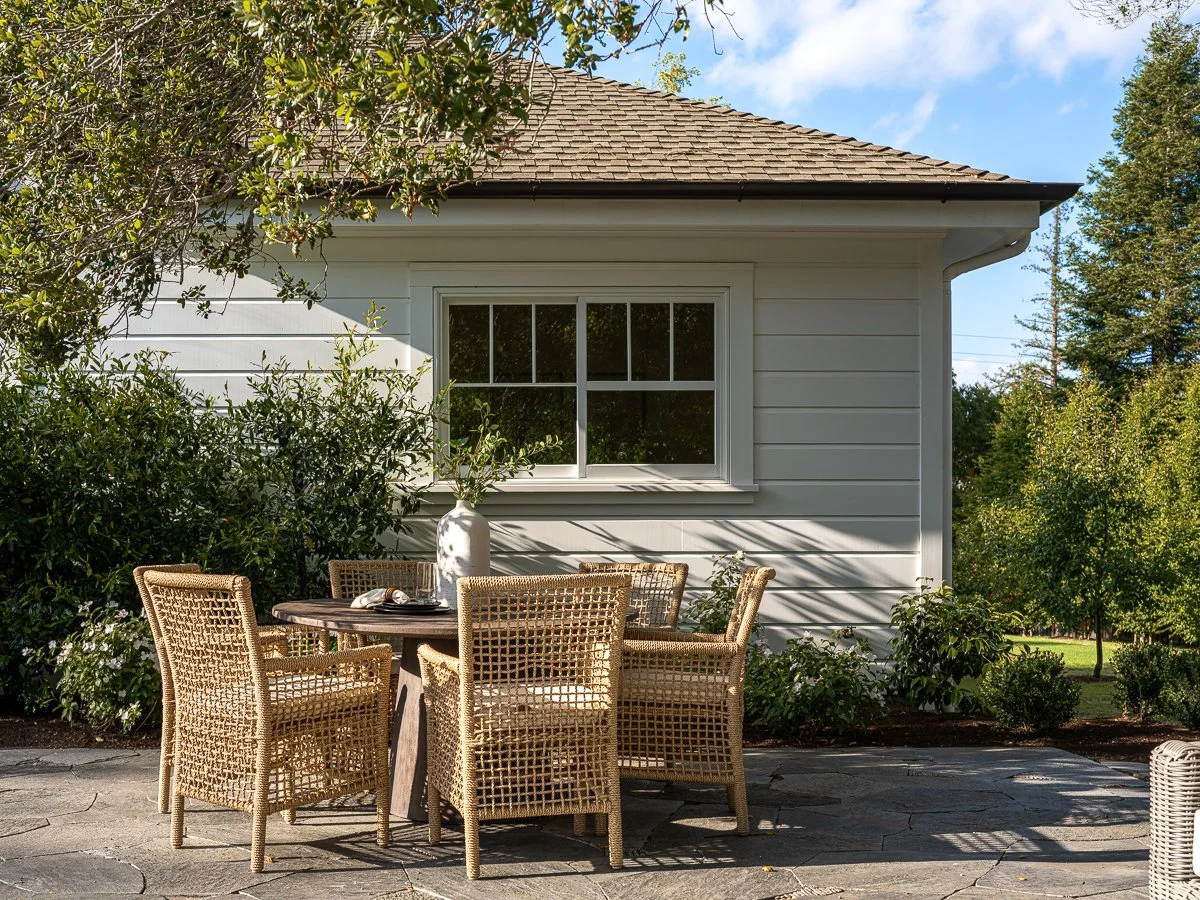 Outdoor patio with a round dining table and wicker chairs in front of a white house with a window, surrounded by green trees and bushes, on a sunny day.