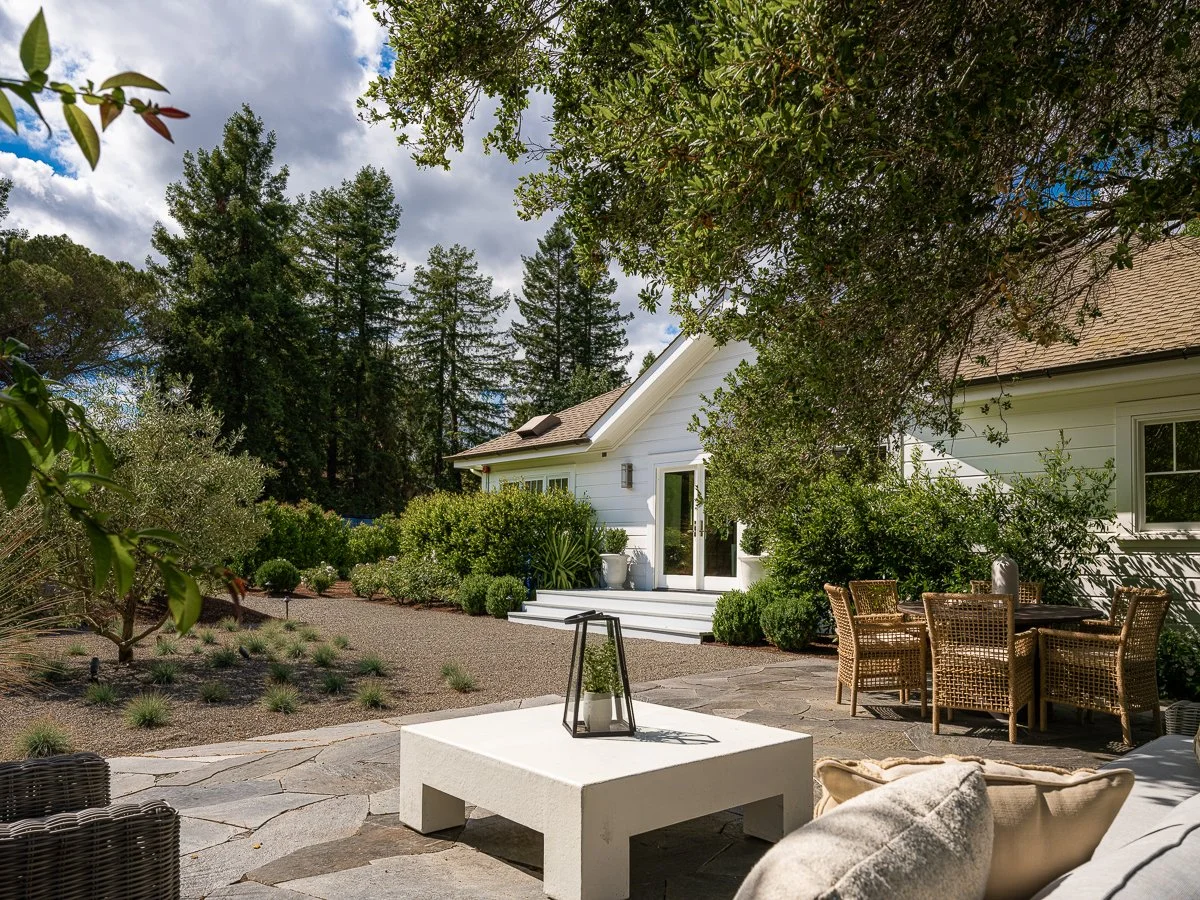A cozy backyard patio with outdoor seating, a white house with steps, surrounded by lush greenery and tall trees under a partly cloudy sky.