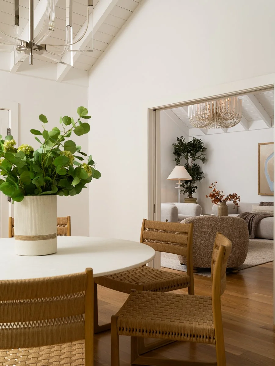 A cozy dining area with a round white table and four wooden chairs, decorated with a large potted green plant. In the background, a living room with a white sofa, a lamp, and a chandelier is visible.