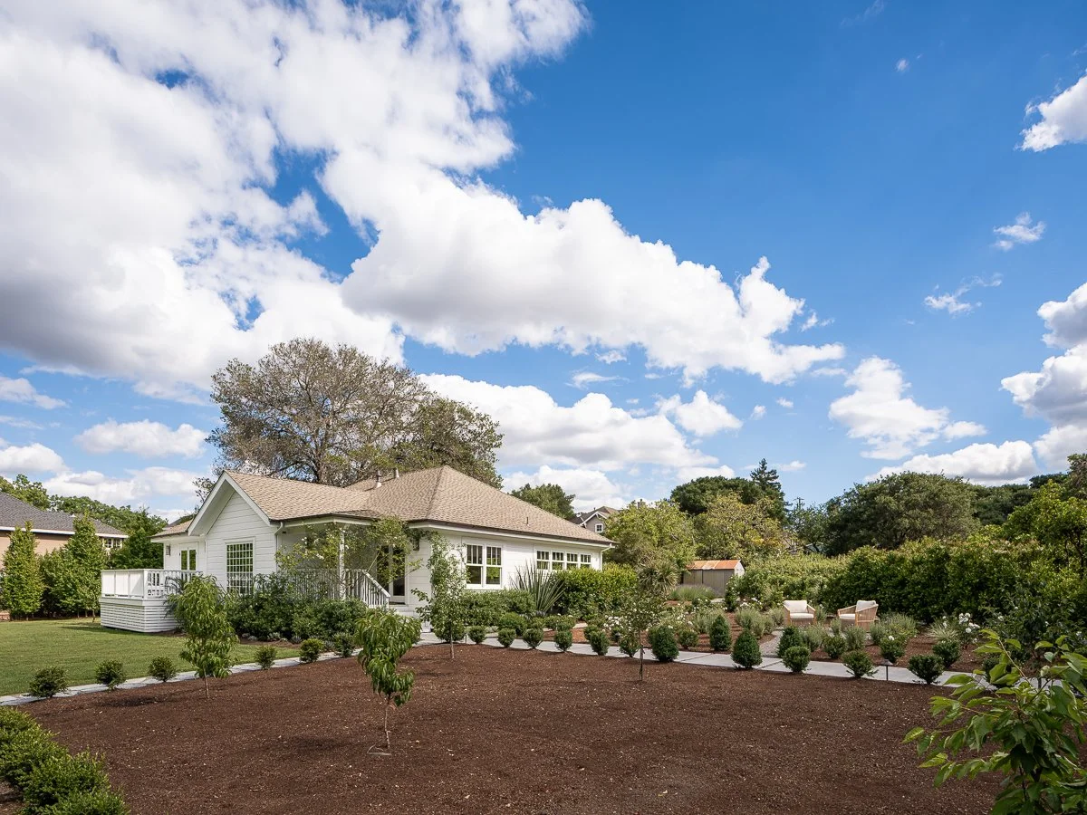 A white house with a porch in a garden with trees and shrubs, under a blue sky with white clouds.