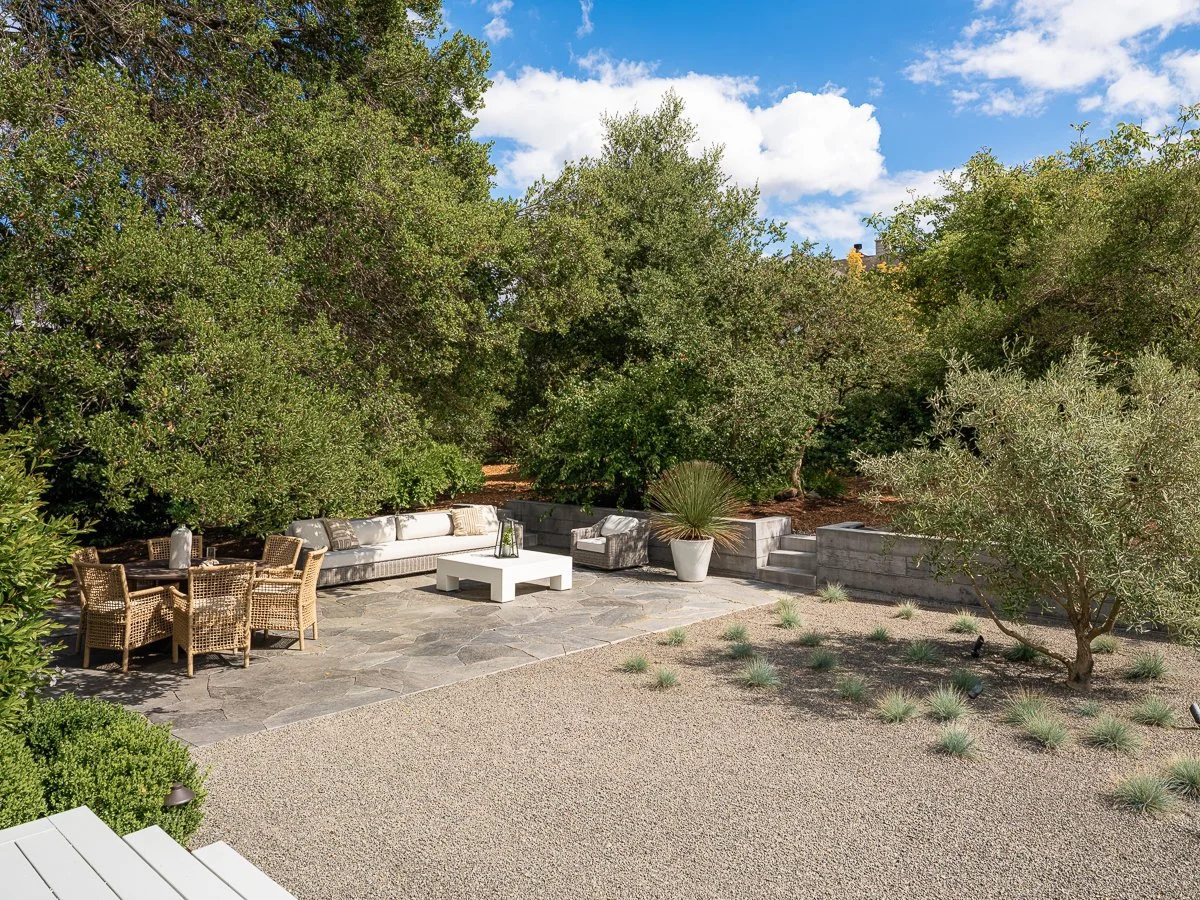 A landscaped backyard patio with outdoor seating, including a white sofa, a wooden dining table with wicker chairs, and potted plants surrounded by trees under a partly cloudy sky.