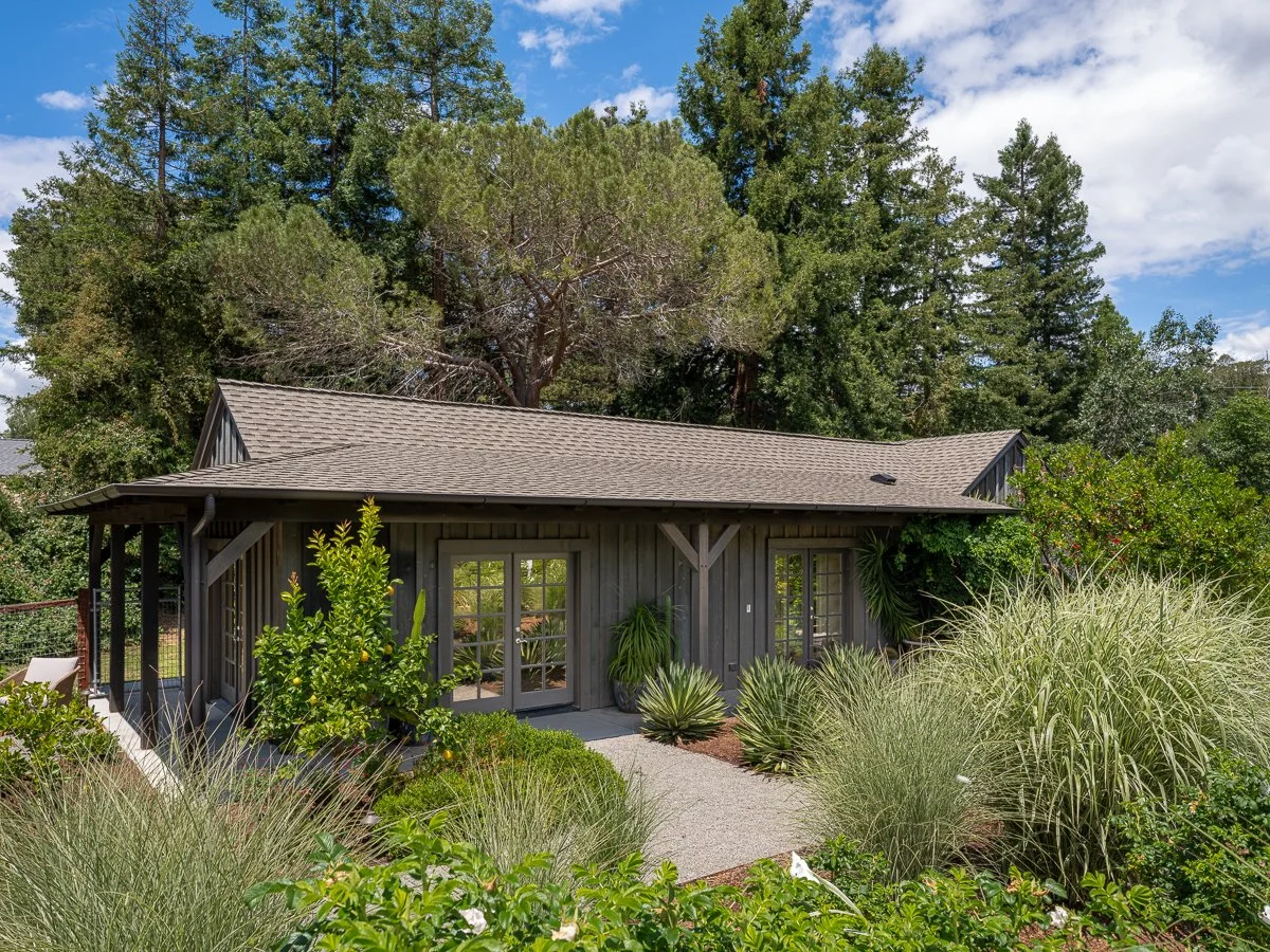 A small, wooden house with a gray roof surrounded by lush green plants and trees under a partly cloudy sky.