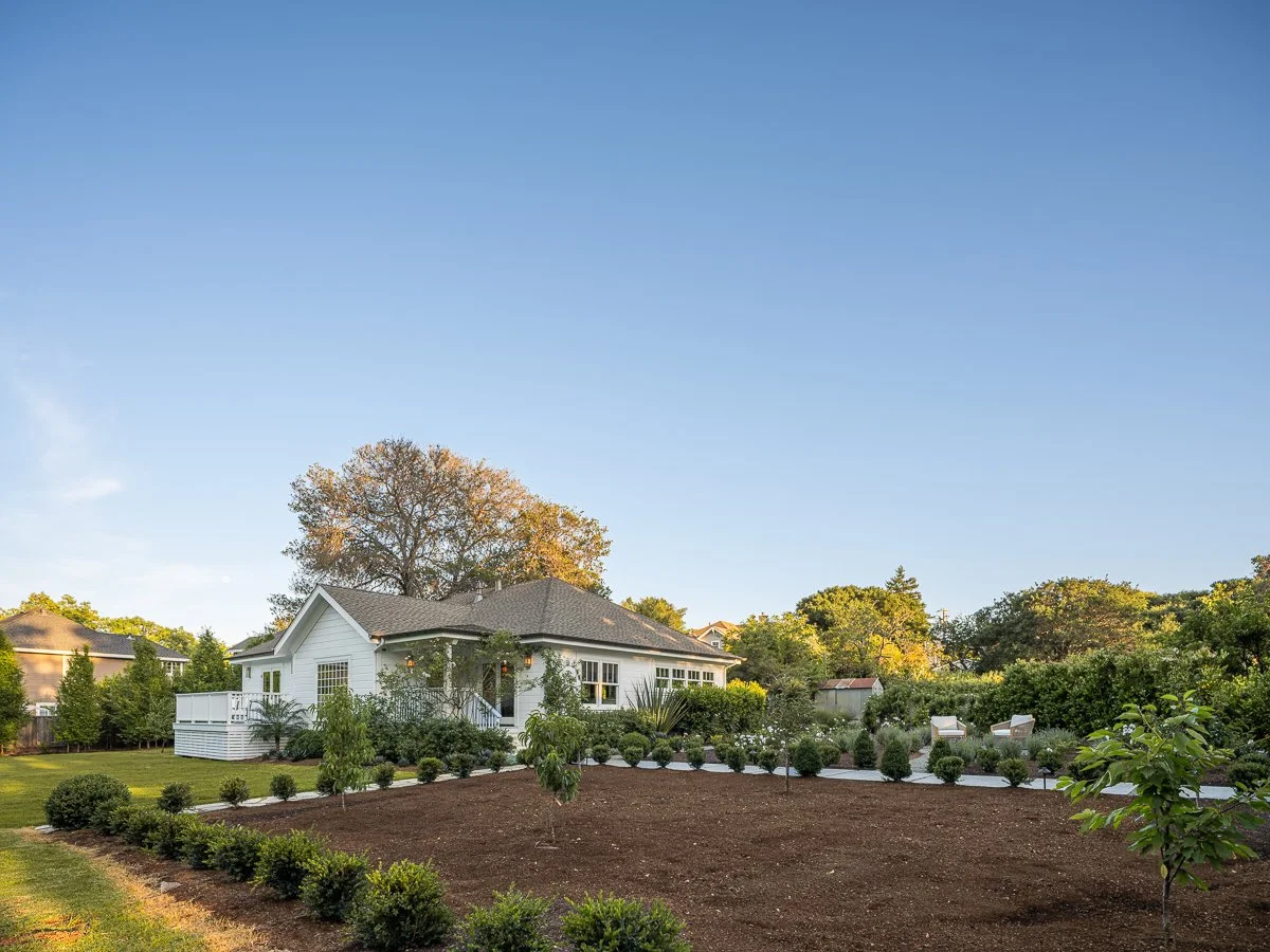 A white house with a gray roof on a large, well-maintained lawn surrounded by trees and shrubs, under a clear blue sky.