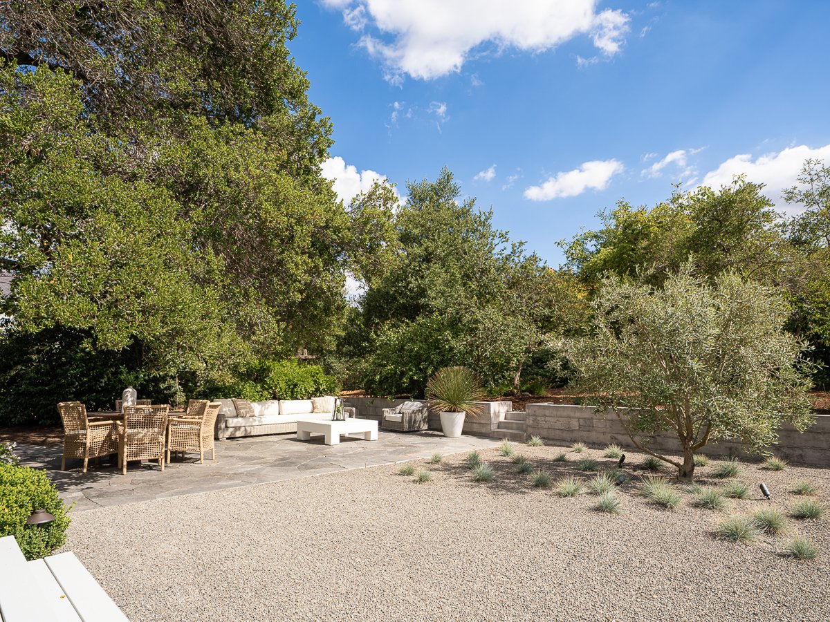 A backyard patio with outdoor furniture, trees, and shrubs under a blue sky with a few clouds.