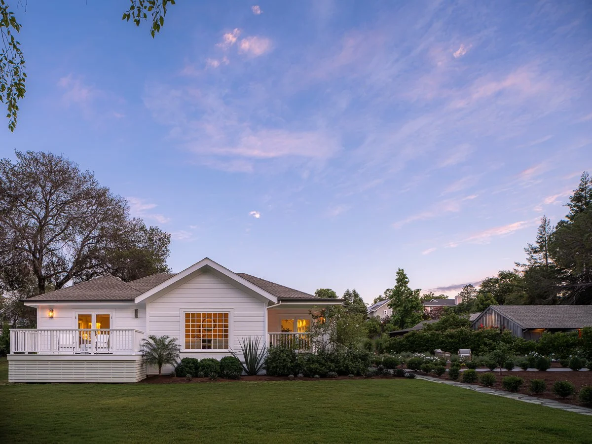 A white house with a deck, outdoor furniture, and garden in the evening, surrounded by trees and a cloudy sky.