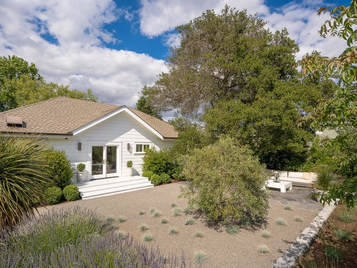 A backyard with a white house, landscaped with bushes, trees, and desert plants, and a sitting area with a bench and pillows under a tree, on a partly cloudy day.