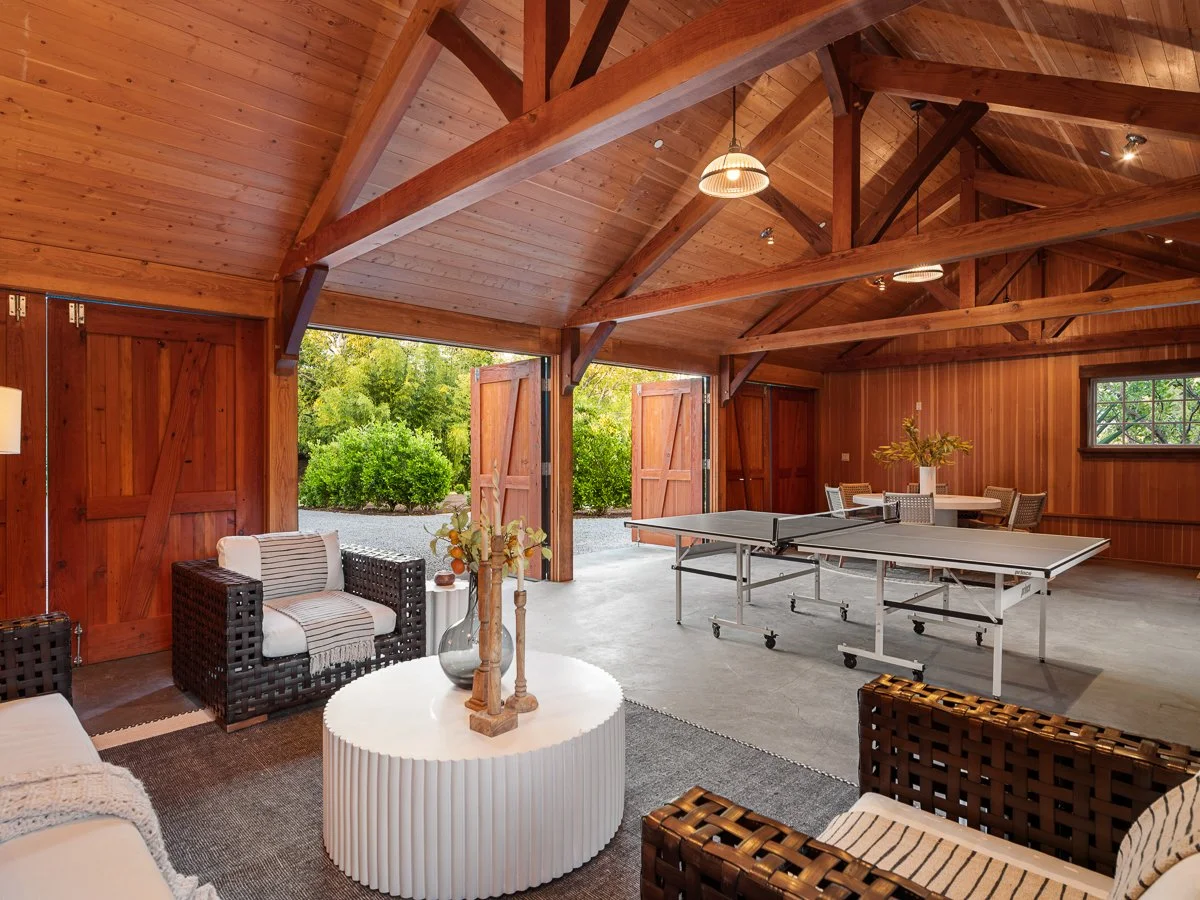 Interior view of a spacious wooden garage or recreation room with open doors, showing outdoor greenery. The room features seating, a coffee table, a ping pong table, and a dining table with chairs.