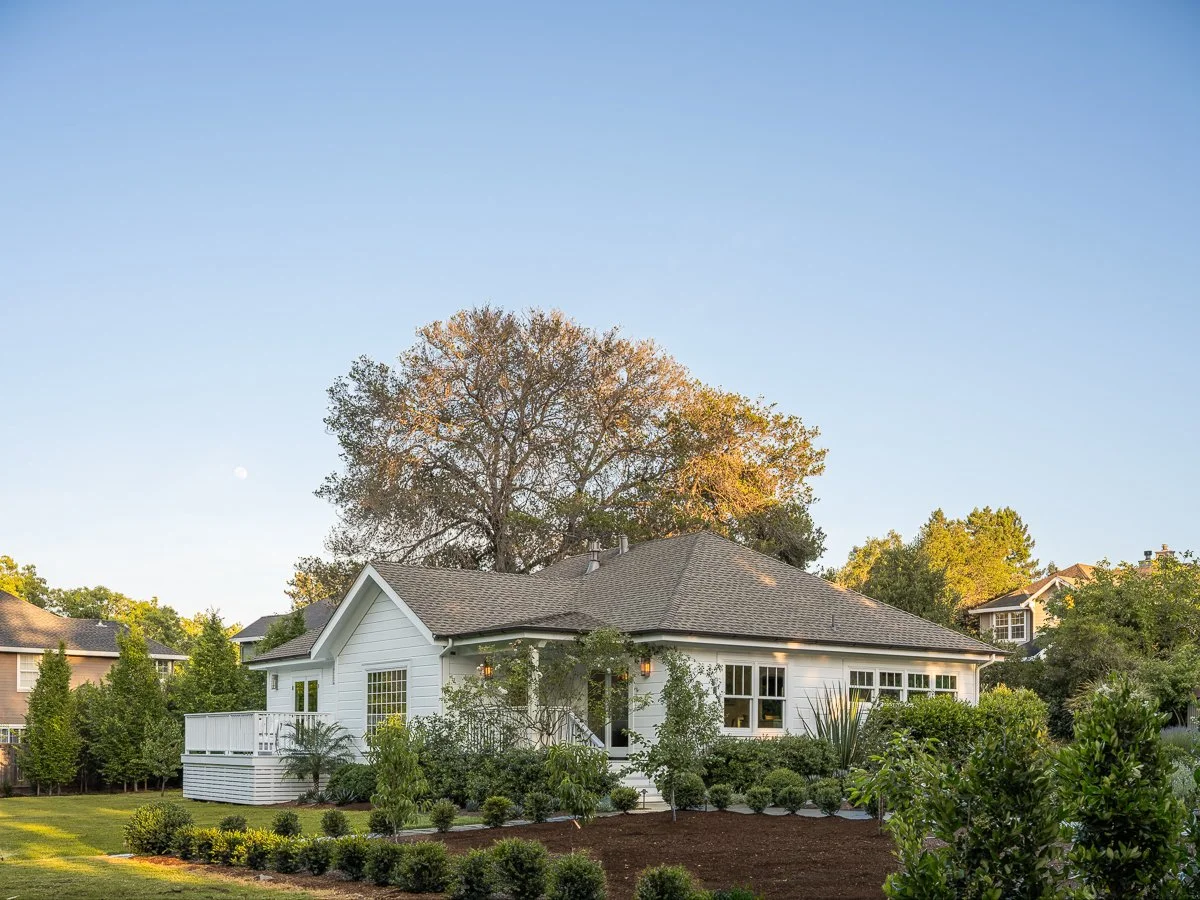 A white house with a gray roof surrounded by lush green shrubs and trees in a suburban neighborhood during daytime.