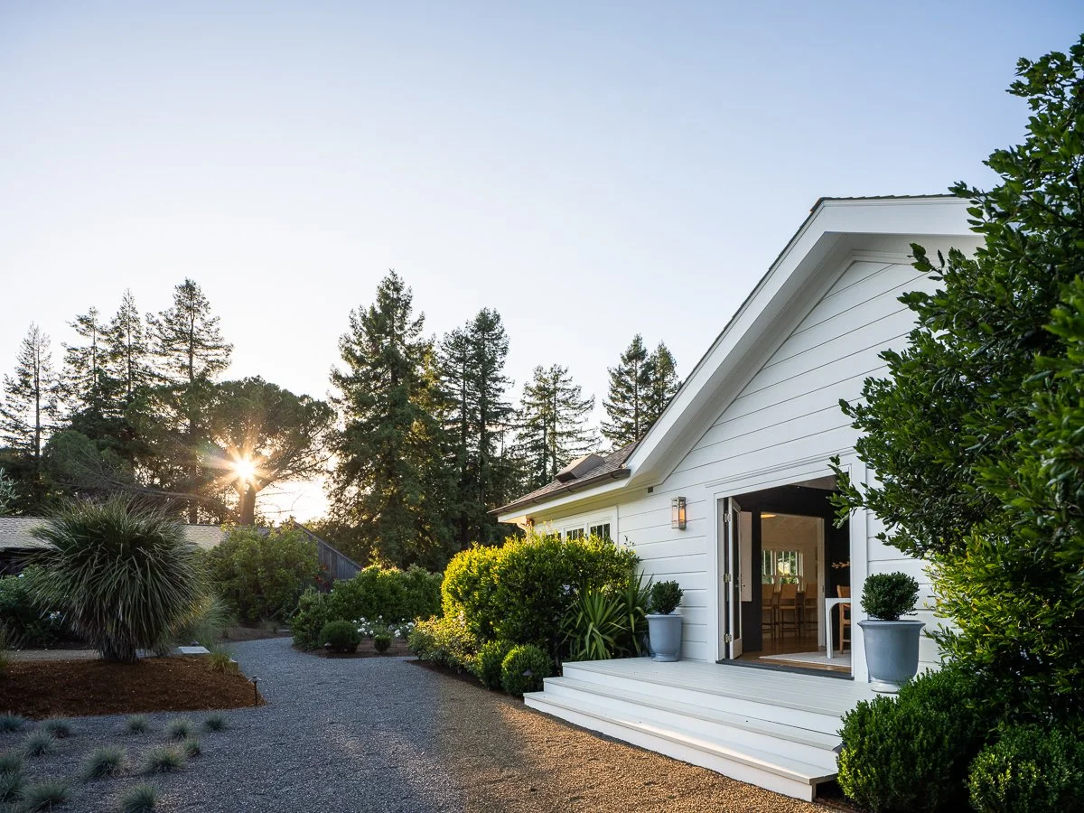 White house with open sliding glass door, surrounded by green bushes and plants, with tall trees and sunlight in the background