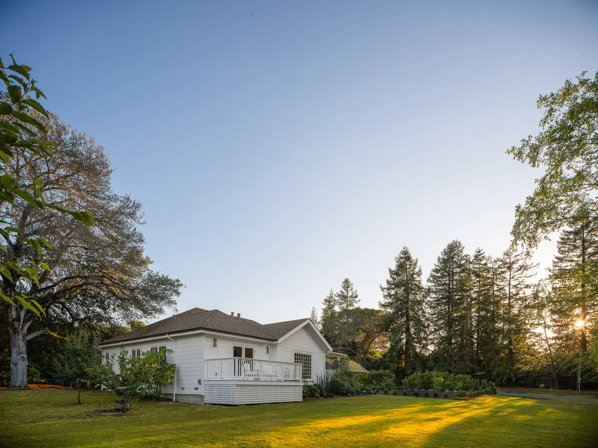 White house with a deck surrounded by green lawn and trees at sunset.
