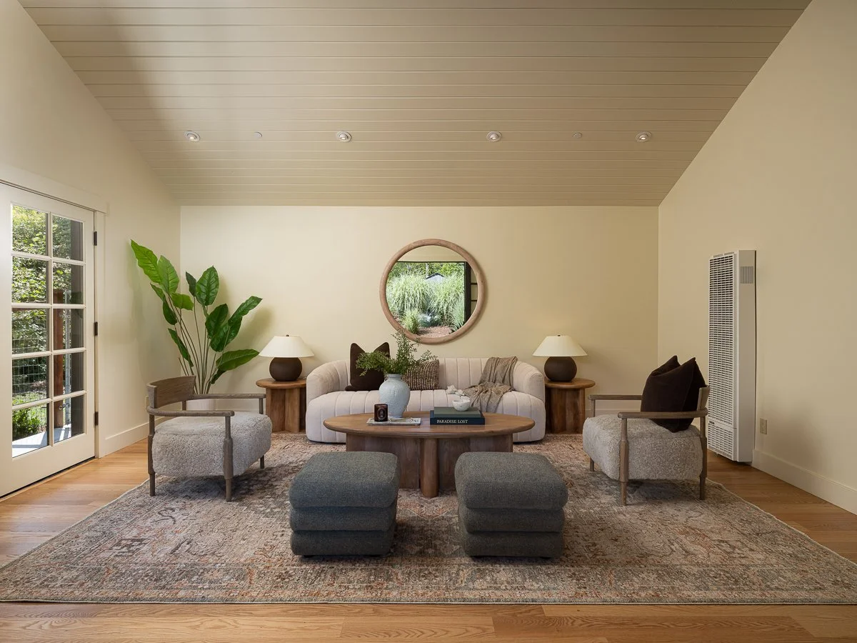 Living room with cream sofa, wooden side tables, armchairs, large potted plant, round mirror, rug, and two ottomans.