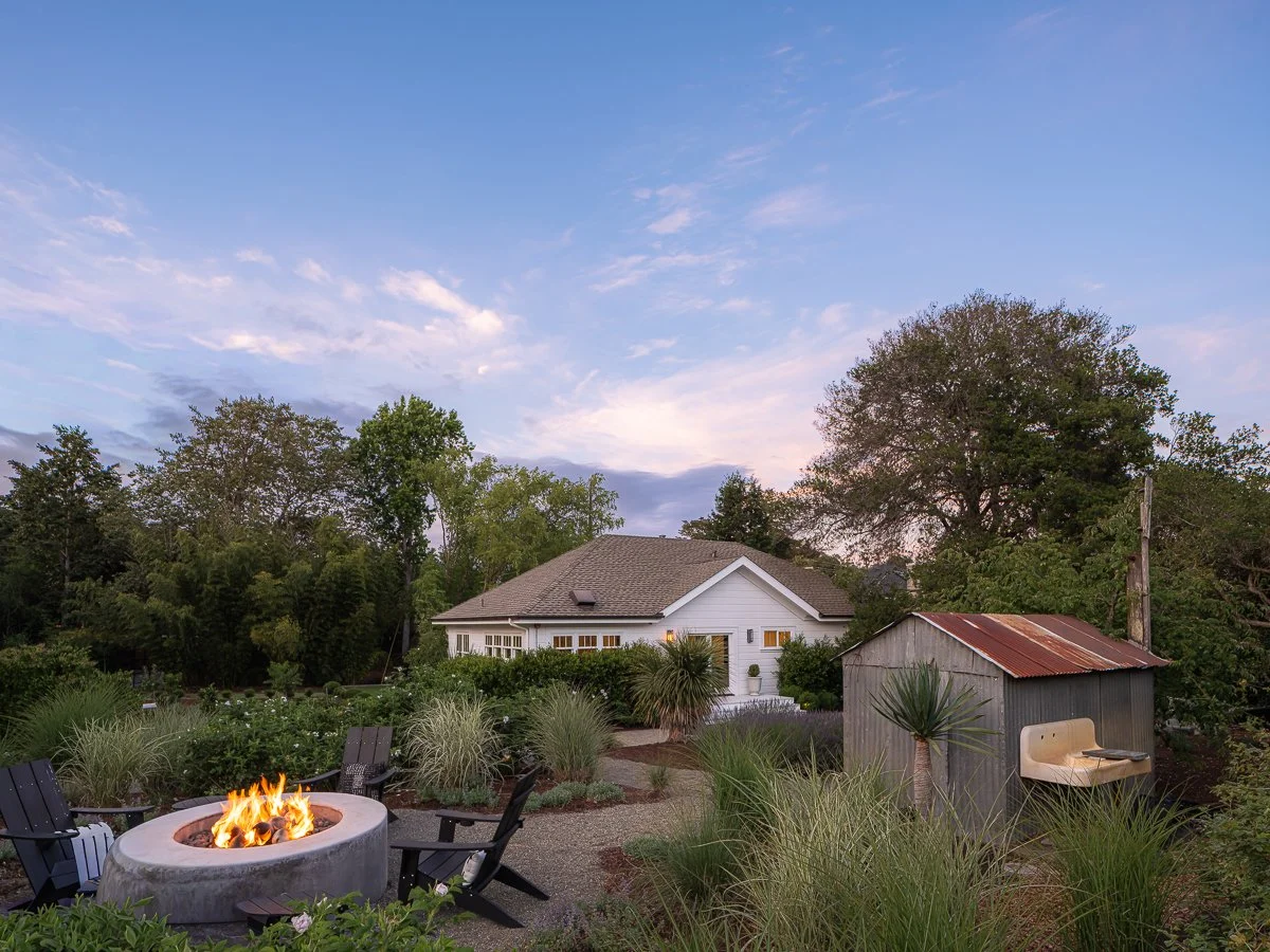 A backyard with a circular fire pit surrounded by chairs, lush landscaping, a white house in the background, and a shed with a rusted metal roof, under a blue sky with some clouds.