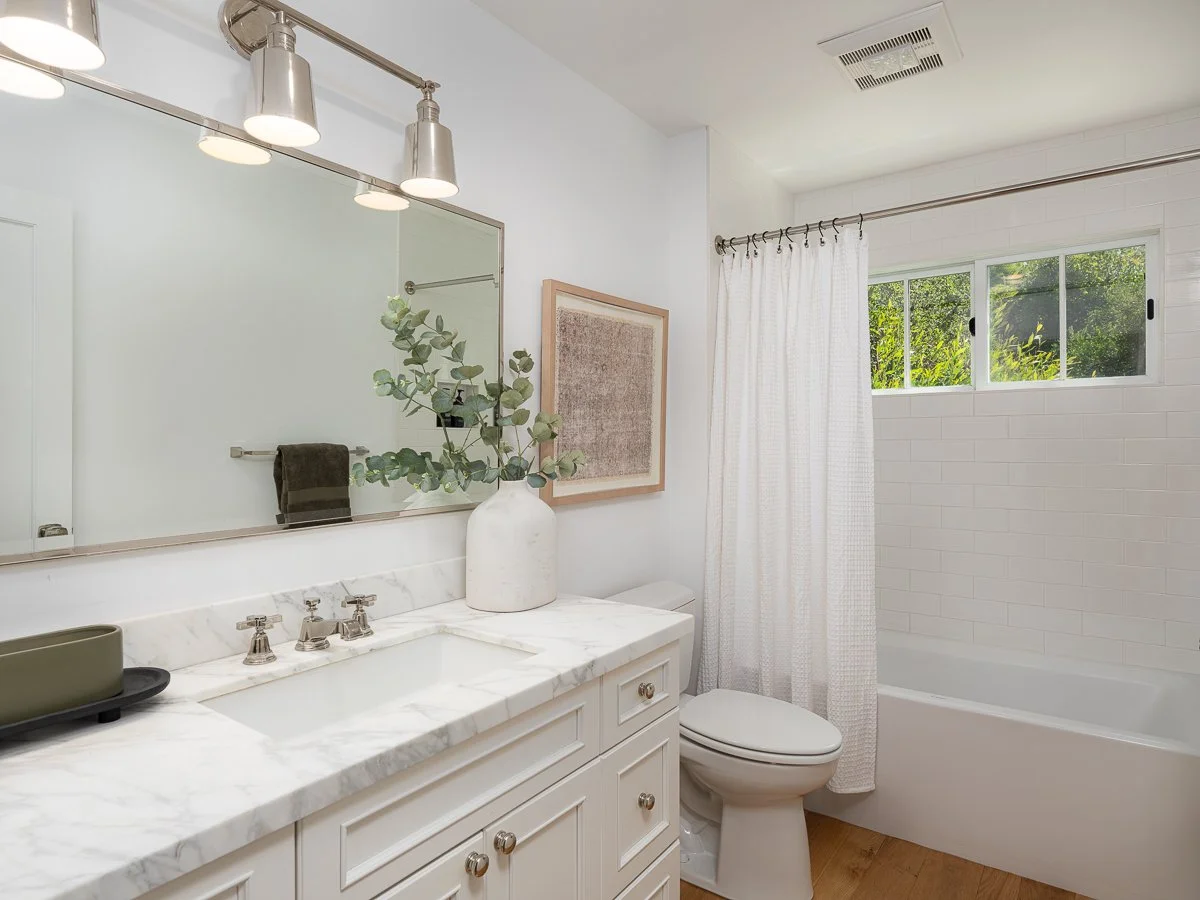 Bathroom with white vanity, marble countertop, large mirror, modern light fixtures, a vase with greenery, framed art, a toilet, window with white curtains, and a bathtub with a white curtain and window with greenery outside.