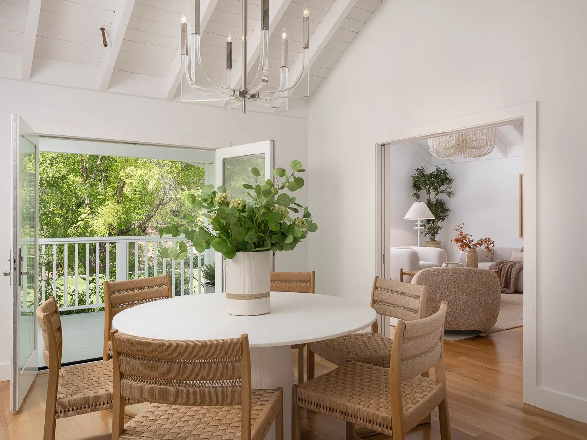 Dining area with white round table, six woven chairs, and a large vase of green foliage in the center. Open double doors lead to a balcony with a view of greenery. A doorway on the right shows a living room with a beige sofa, a white armchair, plants