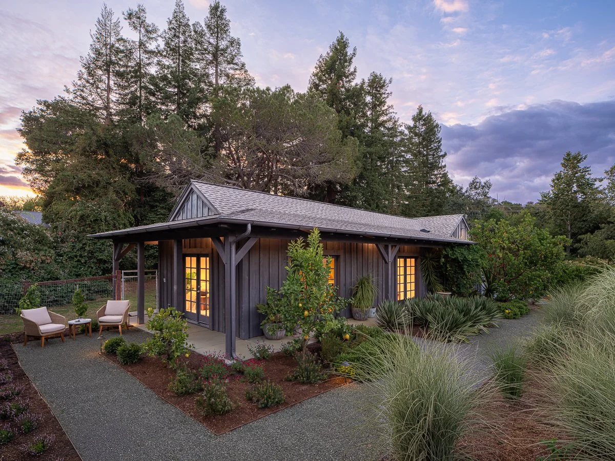 A small dark gray wooden house with large windows glowing from inside, surrounded by a landscaped garden with plants and bushes, two chairs and a small table outside, large trees in the background, and a partly cloudy sky at sunset.