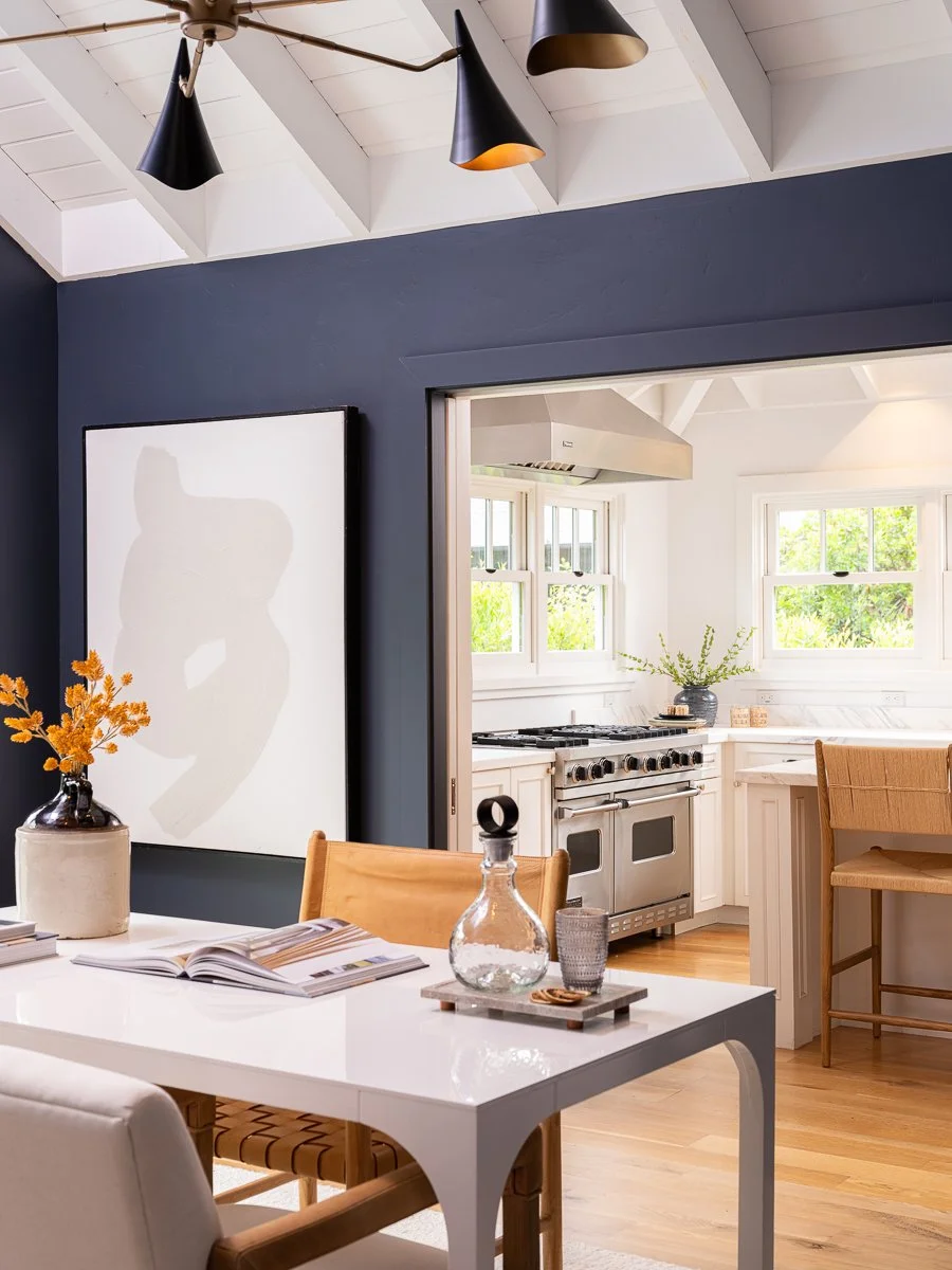 Modern dining area with a white table, wooden chairs, and decorative items, opening into a bright kitchen with white cabinets and windows.