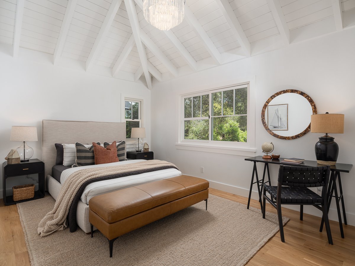 A bright bedroom with a vaulted white ceiling, large windows showing green trees, a bed with a beige headboard, decorative pillows, bedside lamps, a leather bench at the foot of the bed, and a black desk with a mirror and table lamp.