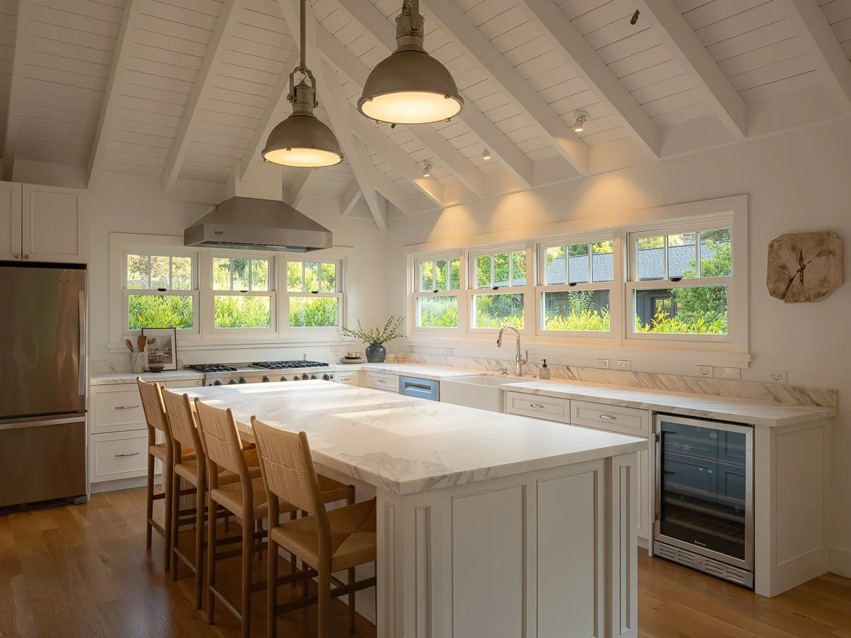 Kitchen with white cabinets, a marble countertop island, stainless steel appliances, multiple windows showing greenery outside, and hanging pendant lights.