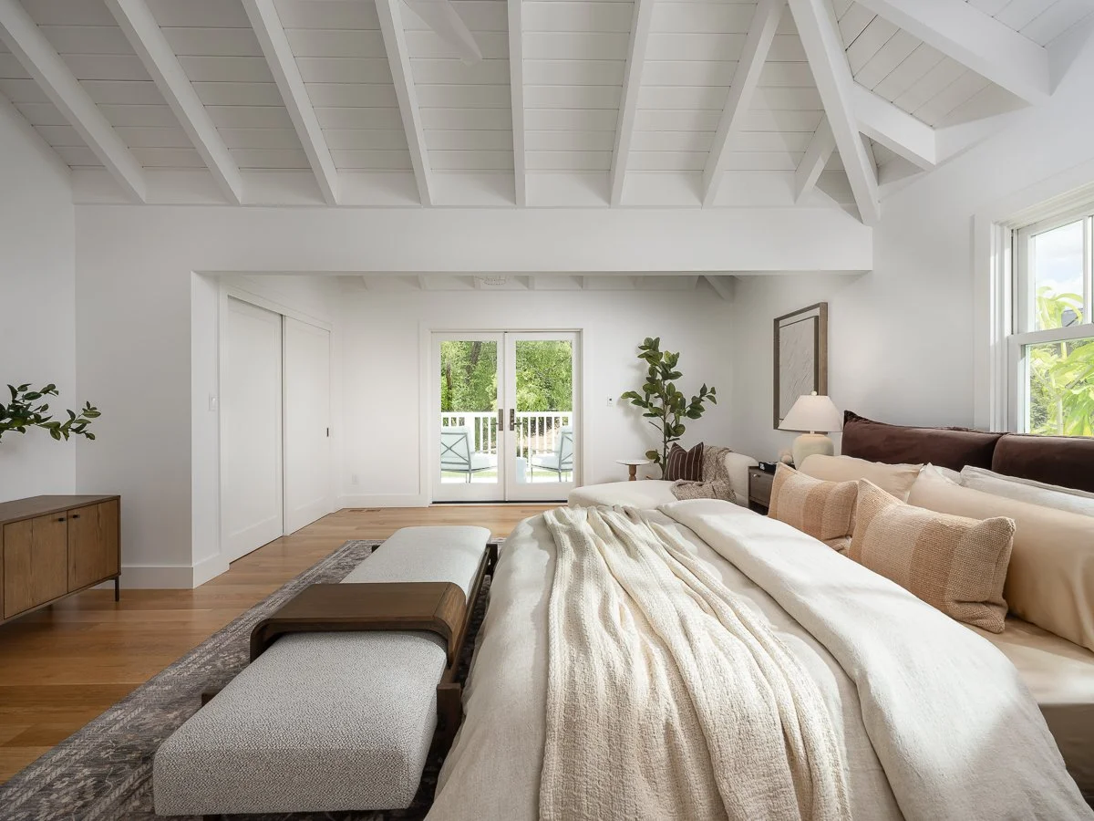 Bright, modern bedroom with white walls and ceiling, bed with beige and white pillows, and small balcony visible through sliding glass door.