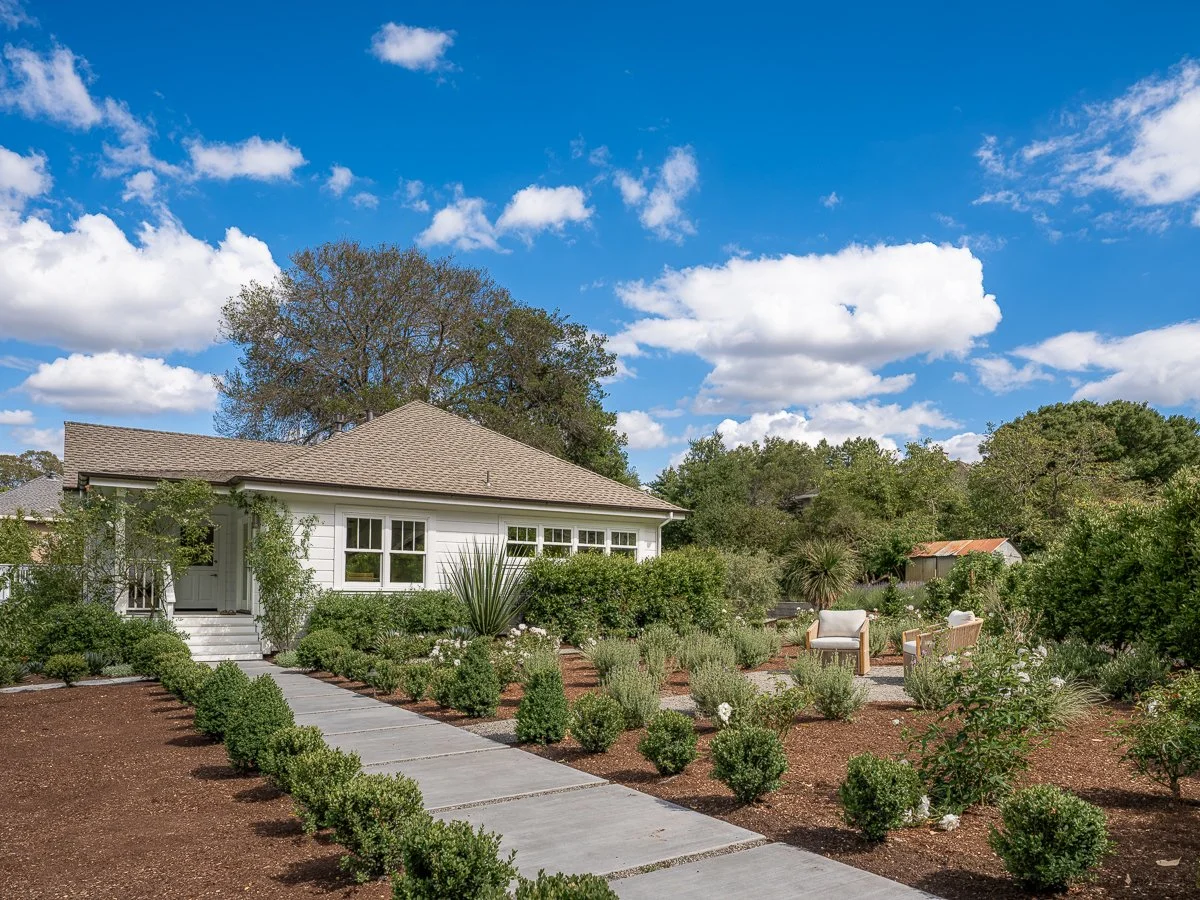A white house with a gray roof surrounded by a green garden with shrubs, plants, and outdoor seating under a partly cloudy blue sky.